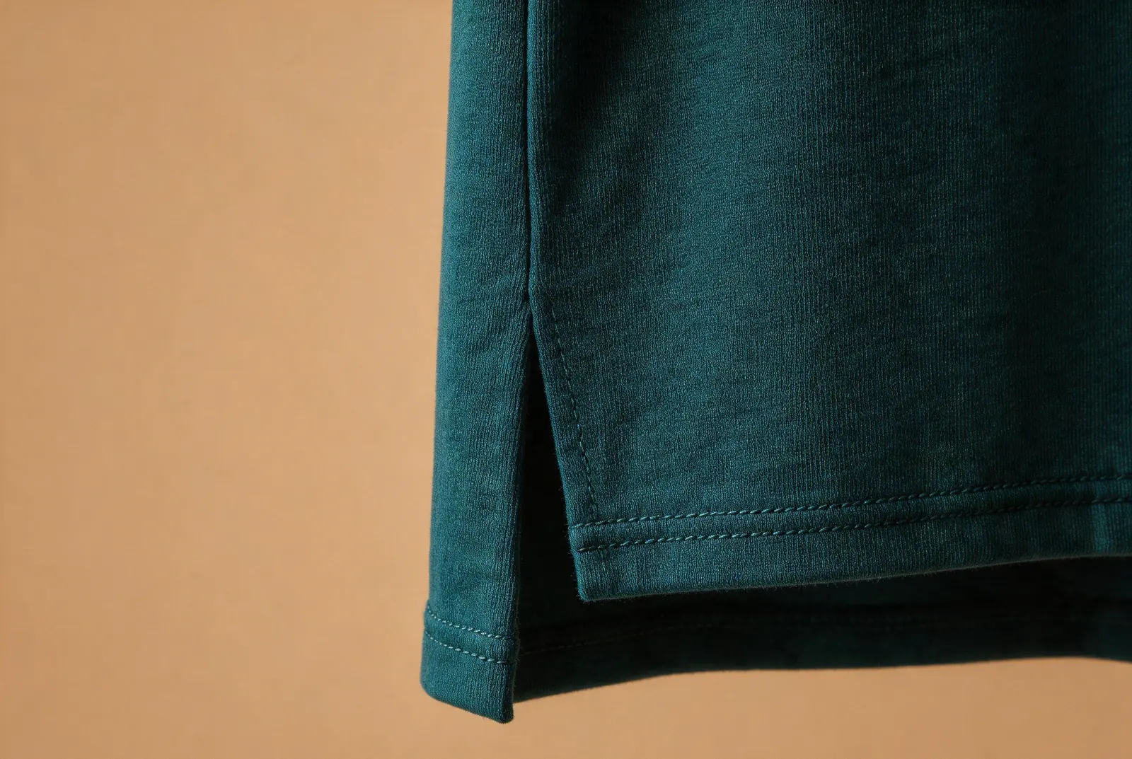 Close-up of a teal T-shirt side opening with two hem edges at different heights against a warm tan background.