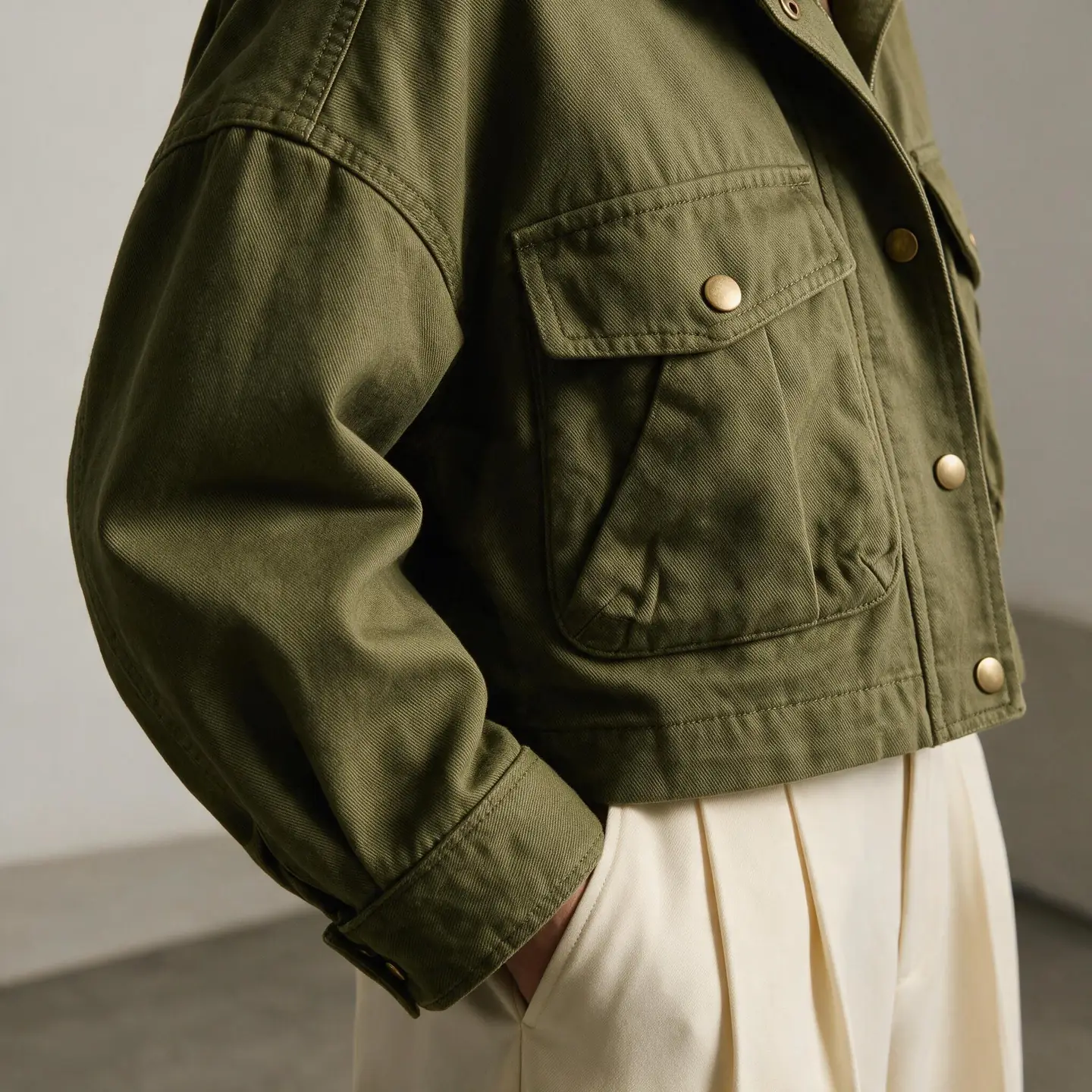 Close-up of an olive utility jacket showing twill texture, shoulder seam, flap pocket, metal snap and cuff detail