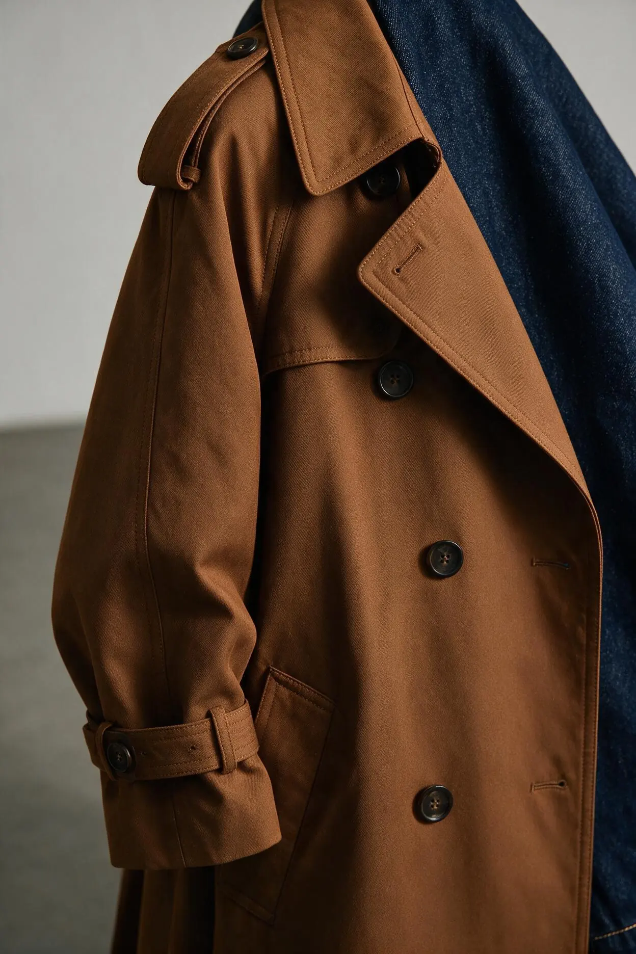 Close-up concept image showing trench coat collar, shoulder tab, buttons, cuff and pocket details beside indigo denim
