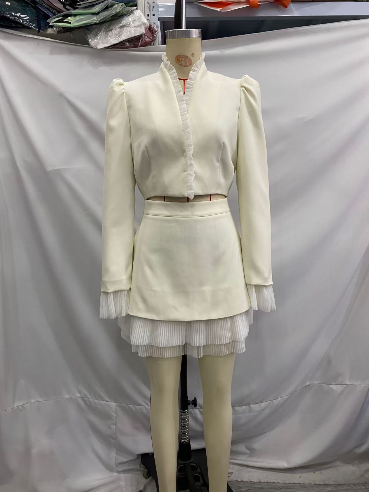 Ivory cropped jacket and short skirt displayed on a dress form, with pleated trim at the neckline, cuffs and skirt hem.