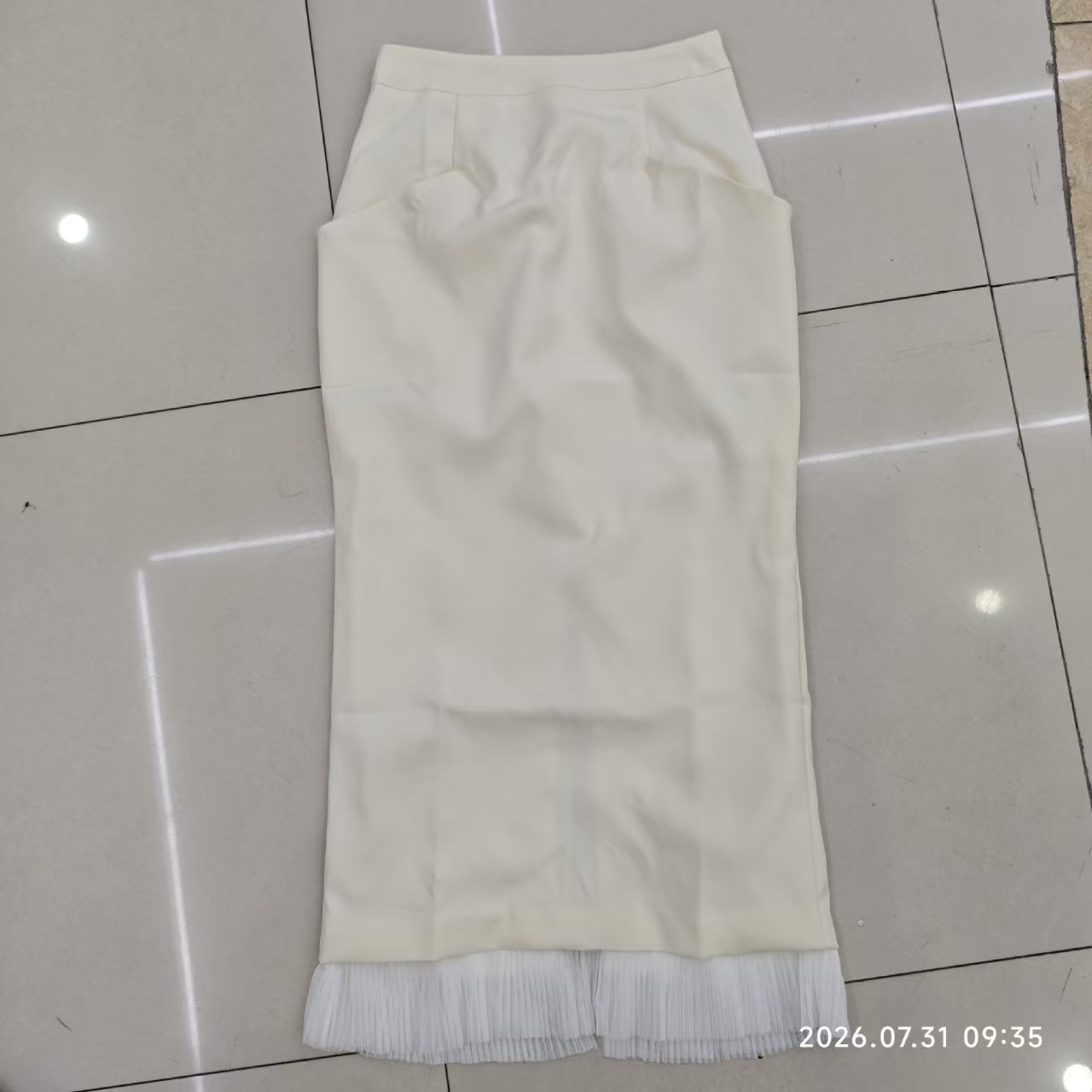 Long ivory skirt laid flat, with shaped pocket openings and pleated hem trim.
