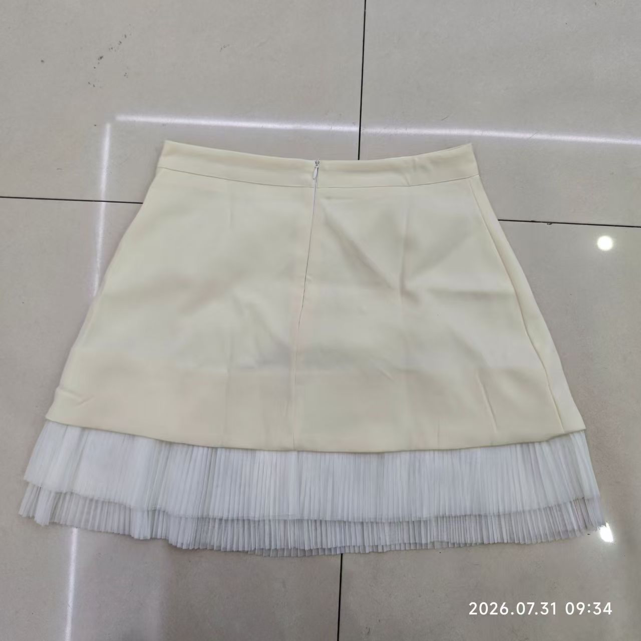Short ivory skirt laid flat, with a waistband, a visible zip and two tiers of pleated hem trim.